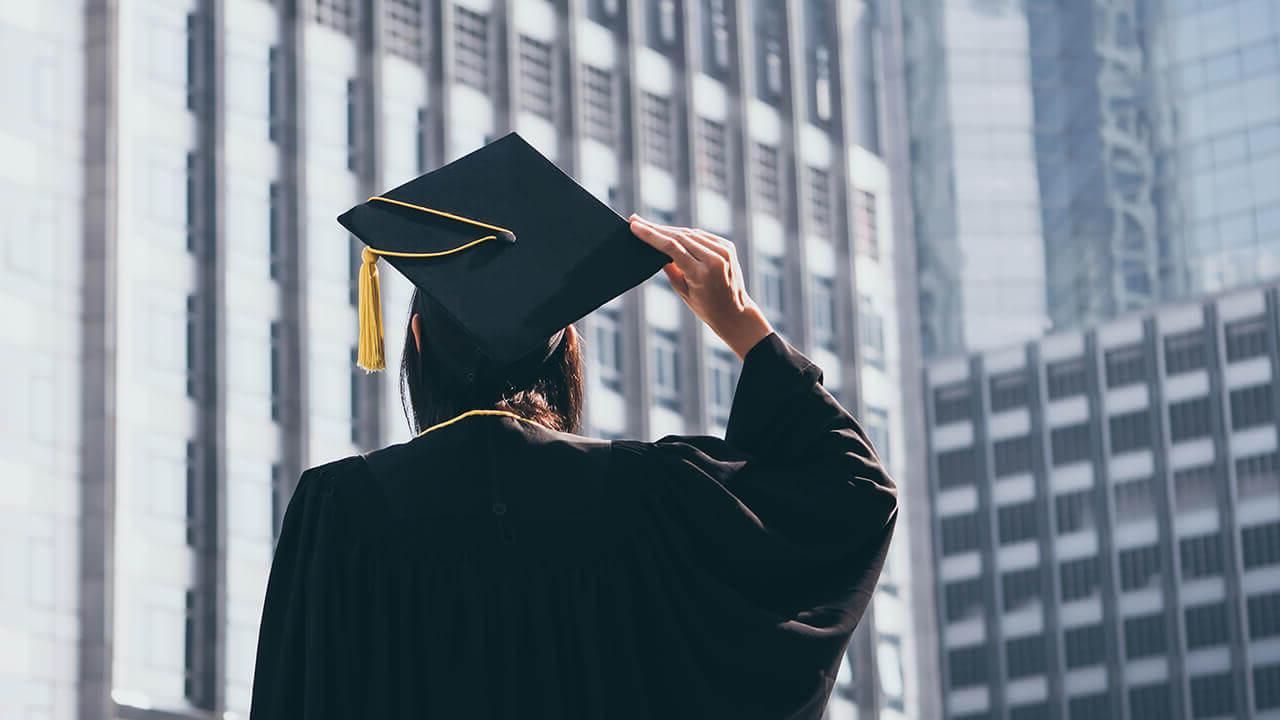 学生 in cap and gown in front of city buildings